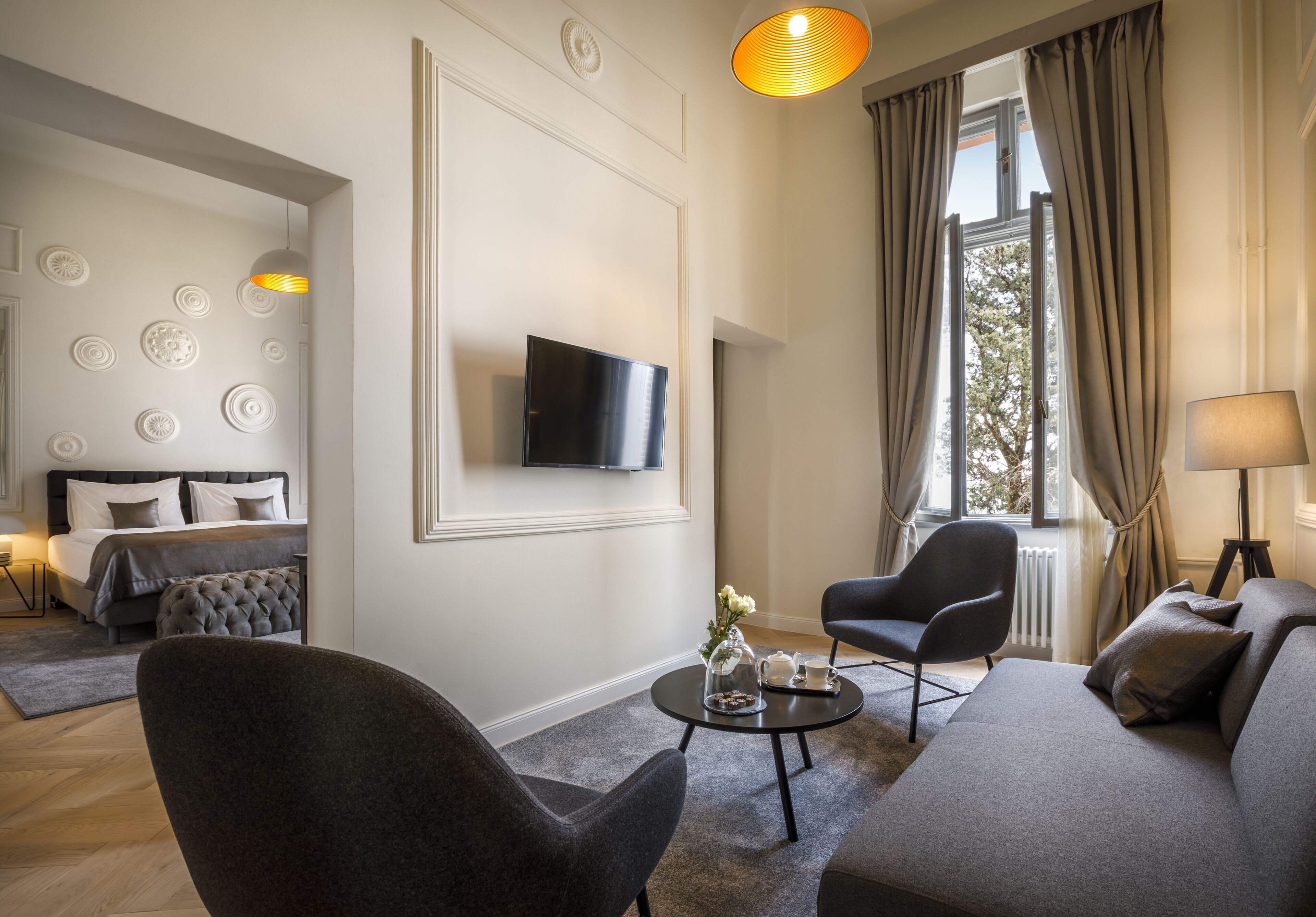 A modern hotel room with a sofa, armchair, and a round coffee table with a vase and teapot. A flat-screen TV is mounted on the wall, and there is a window with curtains.