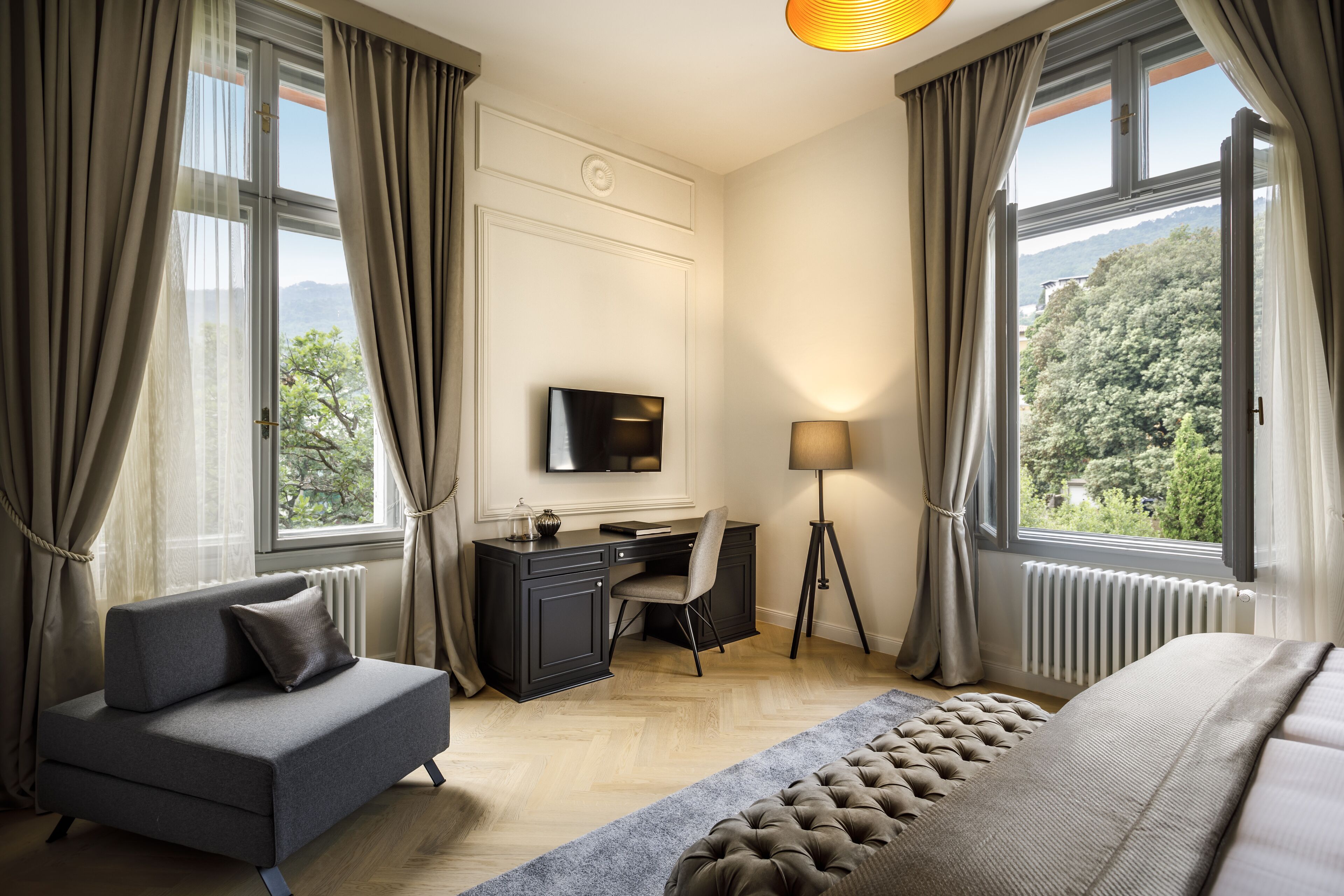 Et moderne soveværelse med sofa, skrivebord, seng og store vinduer med udsigt over grønne omgivelser.