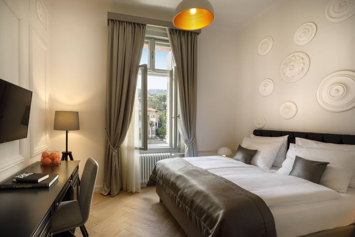 Une chambre d’hôtel avec un lit, un bureau avec une lampe, une chaise, une télévision et une fenêtre avec des rideaux.