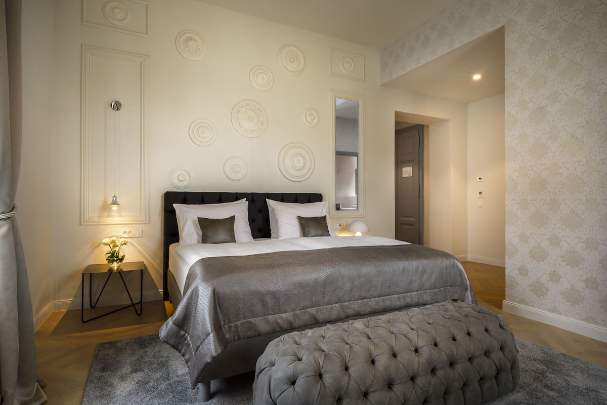 Une chambre à coucher avec un grand lit, une table de chevet avec une lampe, un miroir et un pouf capitonné.
