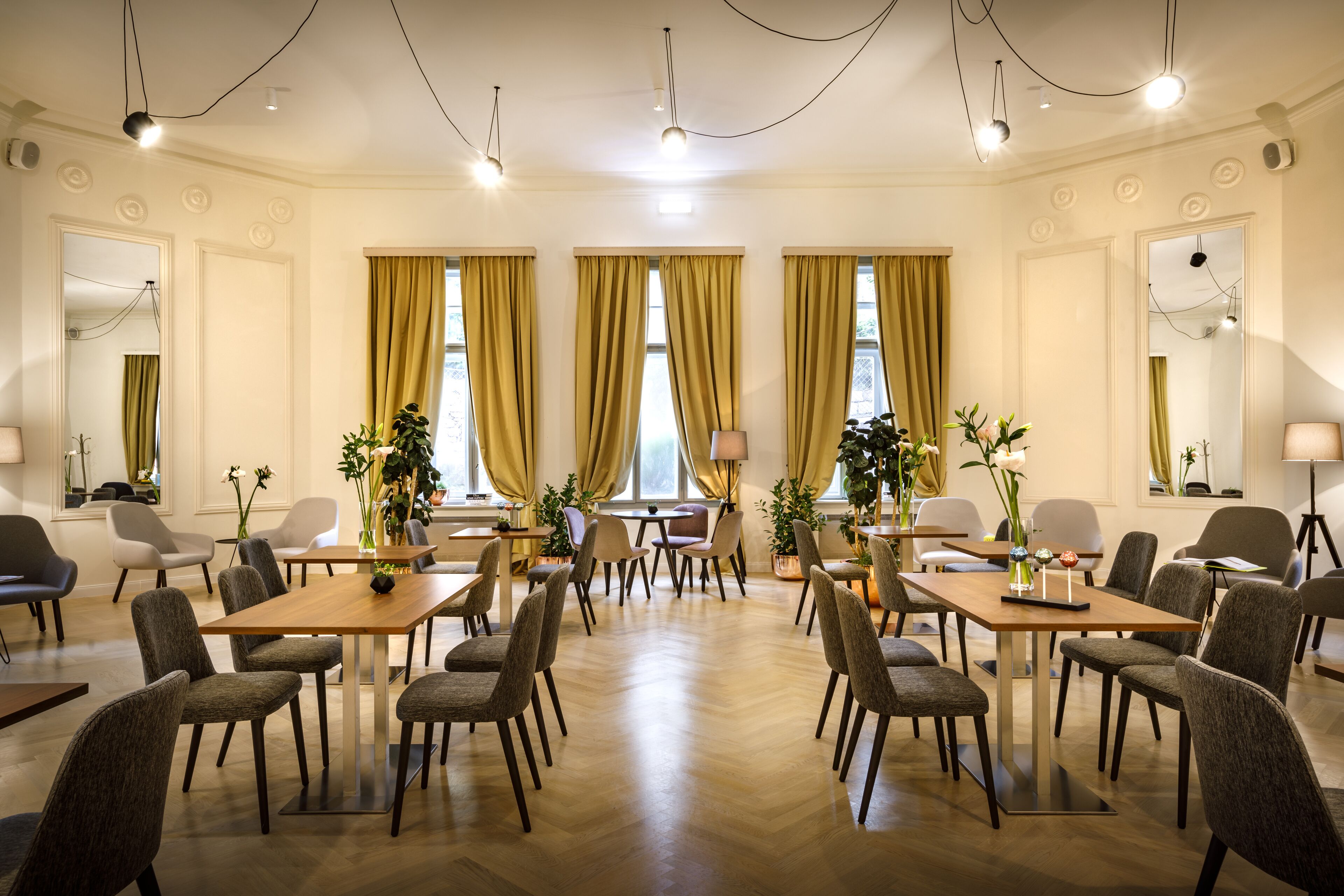 A hotel dining area with multiple tables and chairs, large windows with curtains, and a chandelier.