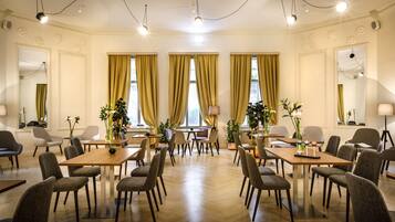 A hotel dining area with multiple tables and chairs, large windows with curtains, and a chandelier.