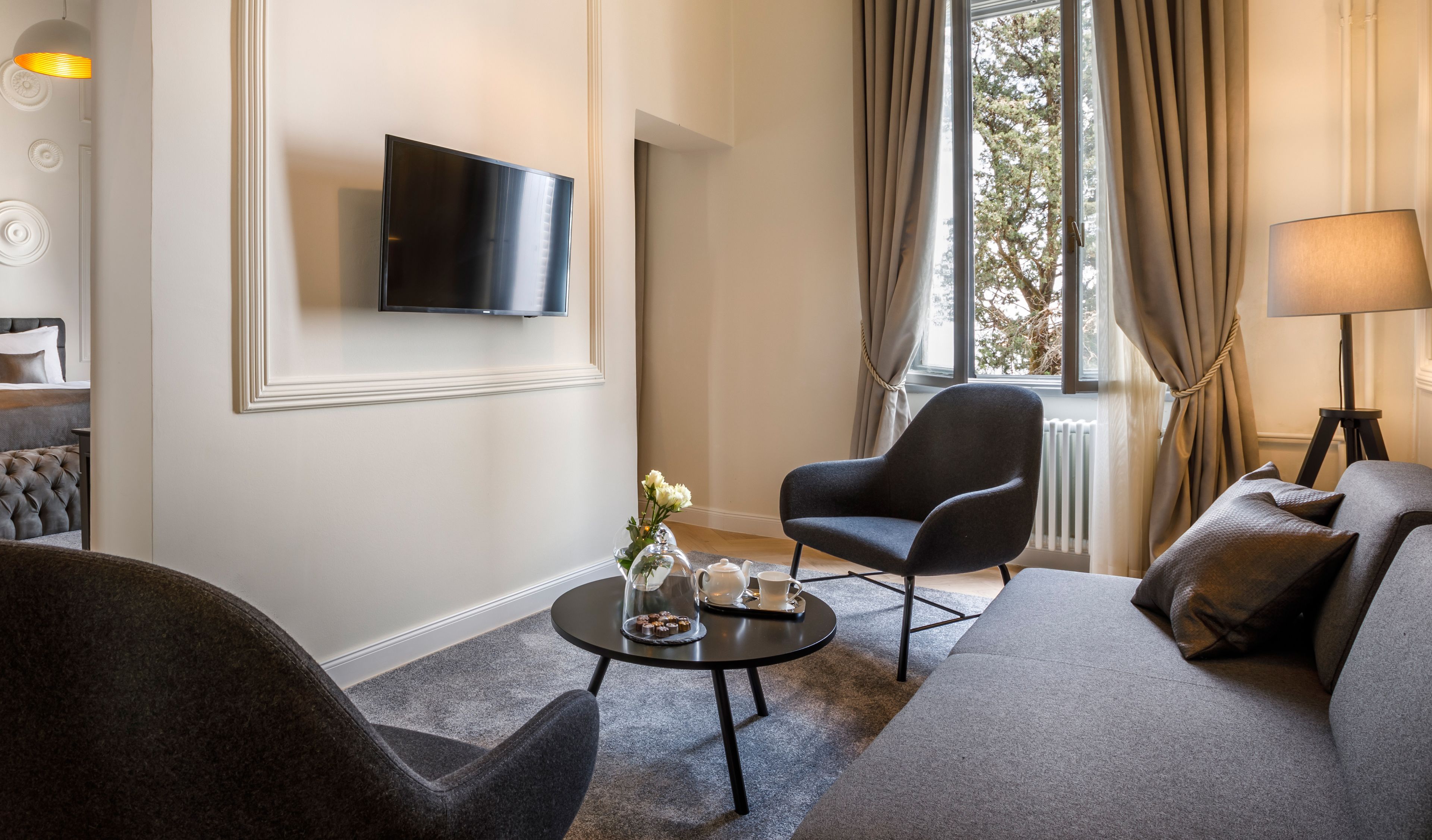 A modern living room with a flat-screen TV mounted on the wall, a comfortable sofa, a dark armchair, and a small round coffee table with a vase of flowers.