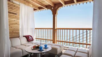 A balcony with a wooden ceiling, a white sofa, a small round table with drinks, and a view of the sea.
