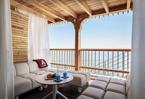 A balcony with a wooden ceiling, a white sofa, a small round table with drinks, and a view of the sea.