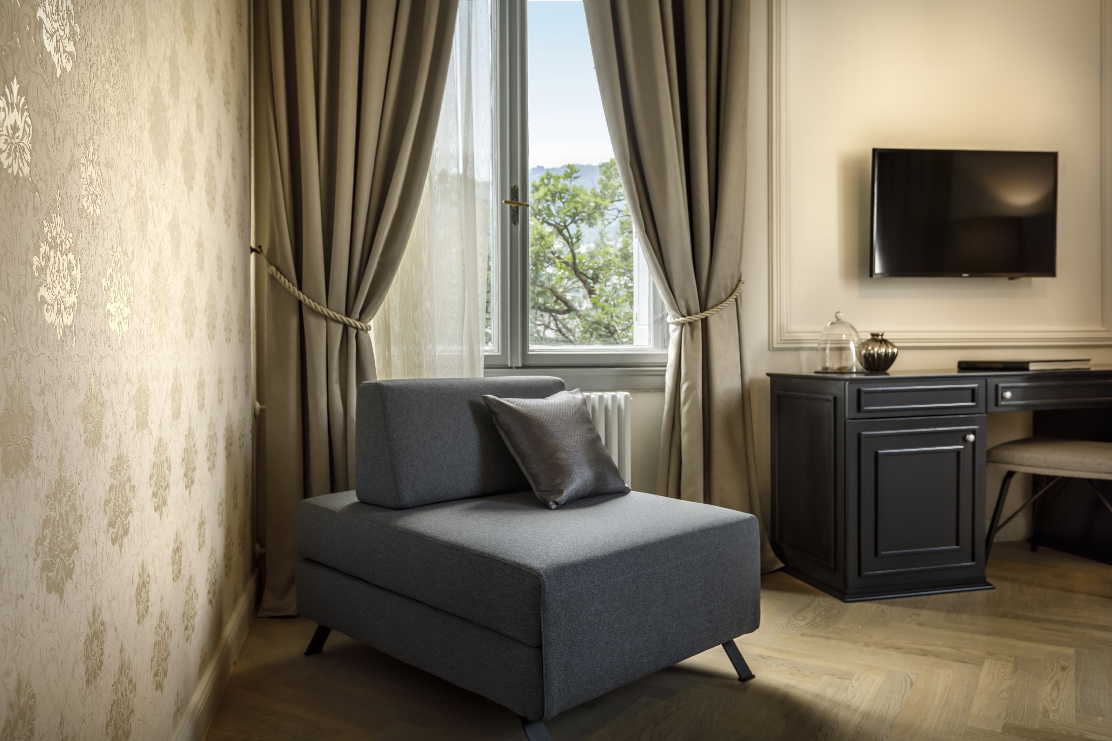 Ein modernes Wohnzimmer mit grauem Sofa, einem Flachbildfernseher an der Wand und Blick ins Freie durch ein fensterförmiges Fenster mit Vorhängen.