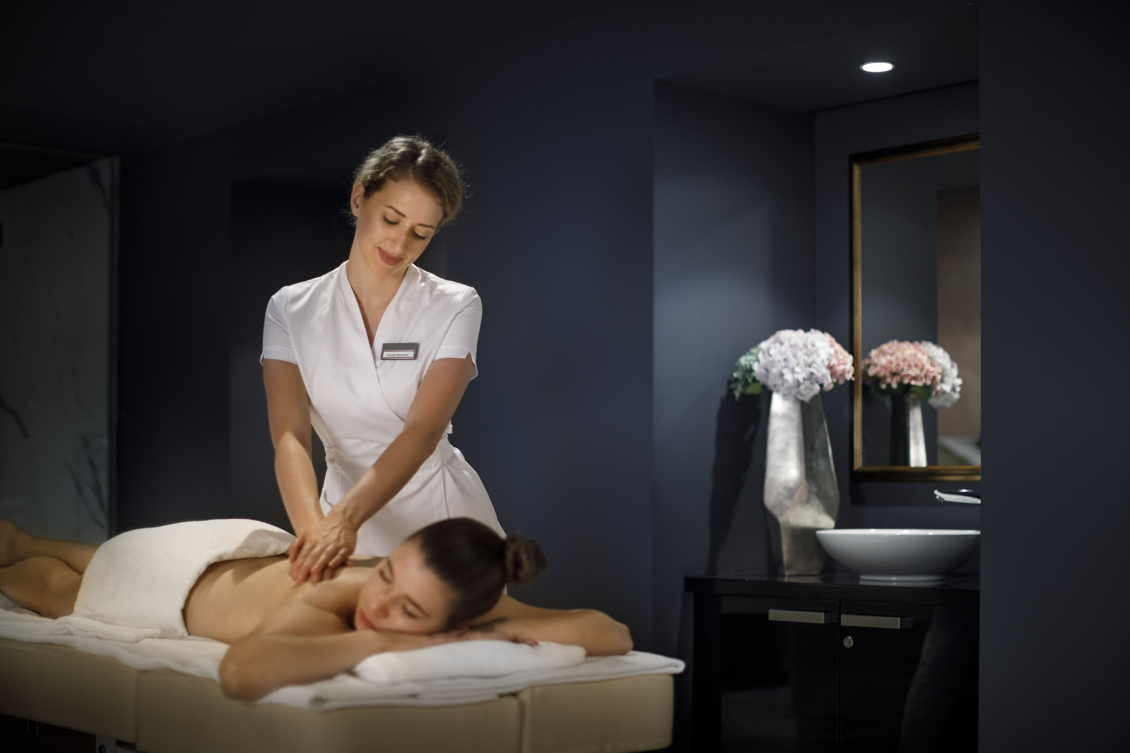 A massage therapy session in a dimly lit room with a dark wall, a mirror, and a vase with flowers.