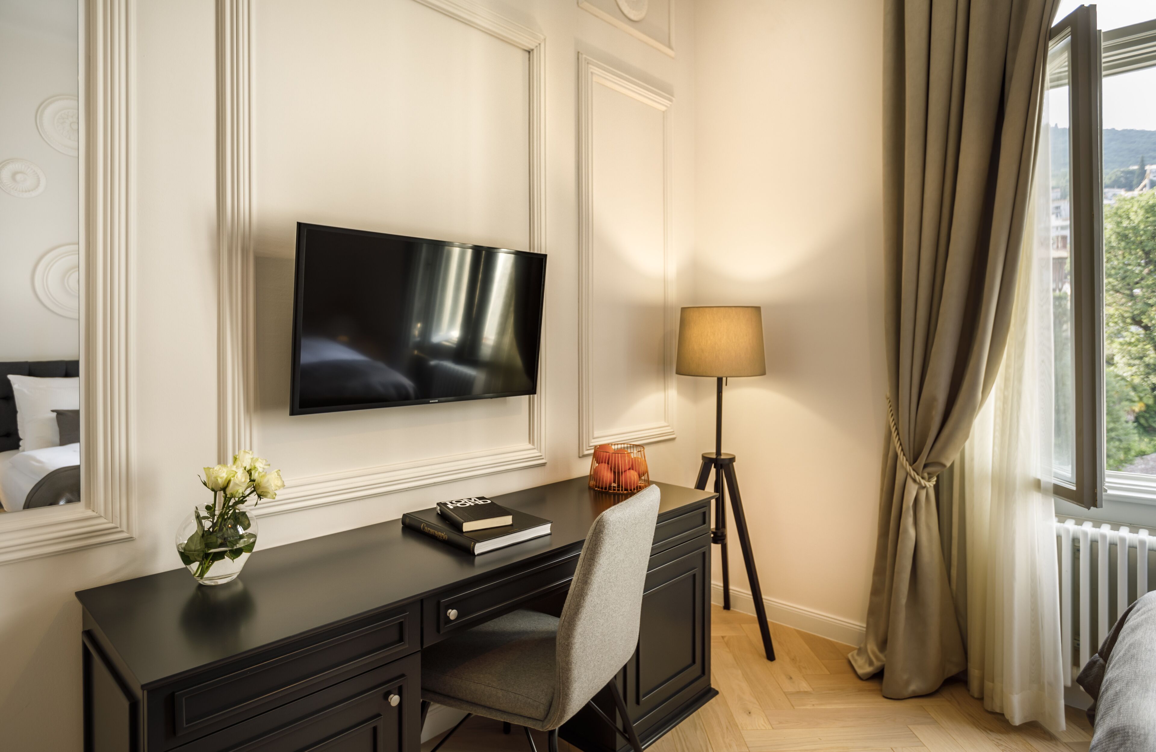 A hotel room with a flat-screen TV mounted on the wall, a desk with a lamp, a chair, and a window with curtains.