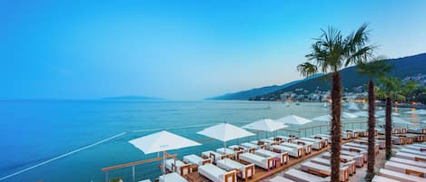 A seaside resort with sun loungers and umbrellas, a wooden deck, and a view of the sea and distant hills.