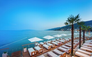 A seaside resort with sun loungers and umbrellas, a wooden deck, and a view of the sea and distant hills.