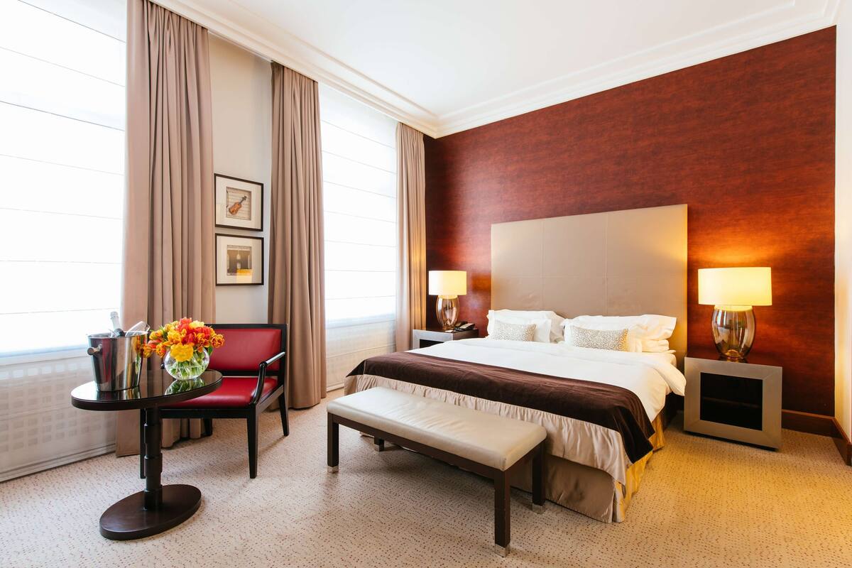 A hotel room with a large bed, bedside lamps, a red chair, a small table with a vase of flowers, and a bench.