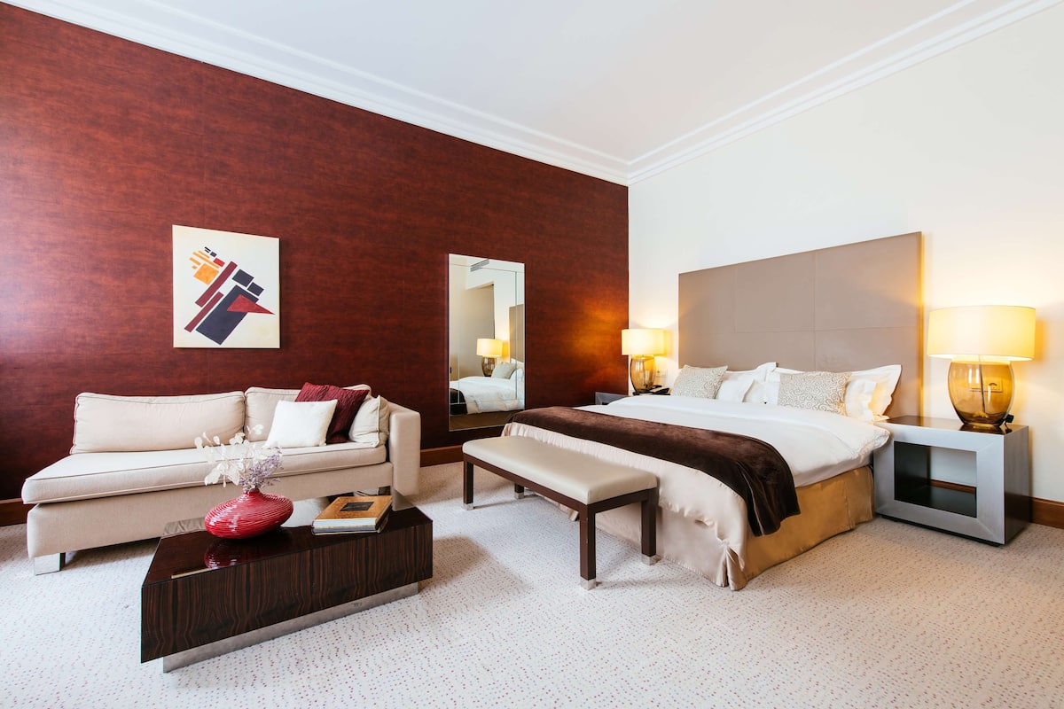 A hotel room with a bed, a sofa, a bench, a coffee table, a bedside table with lamps, and a mirror.