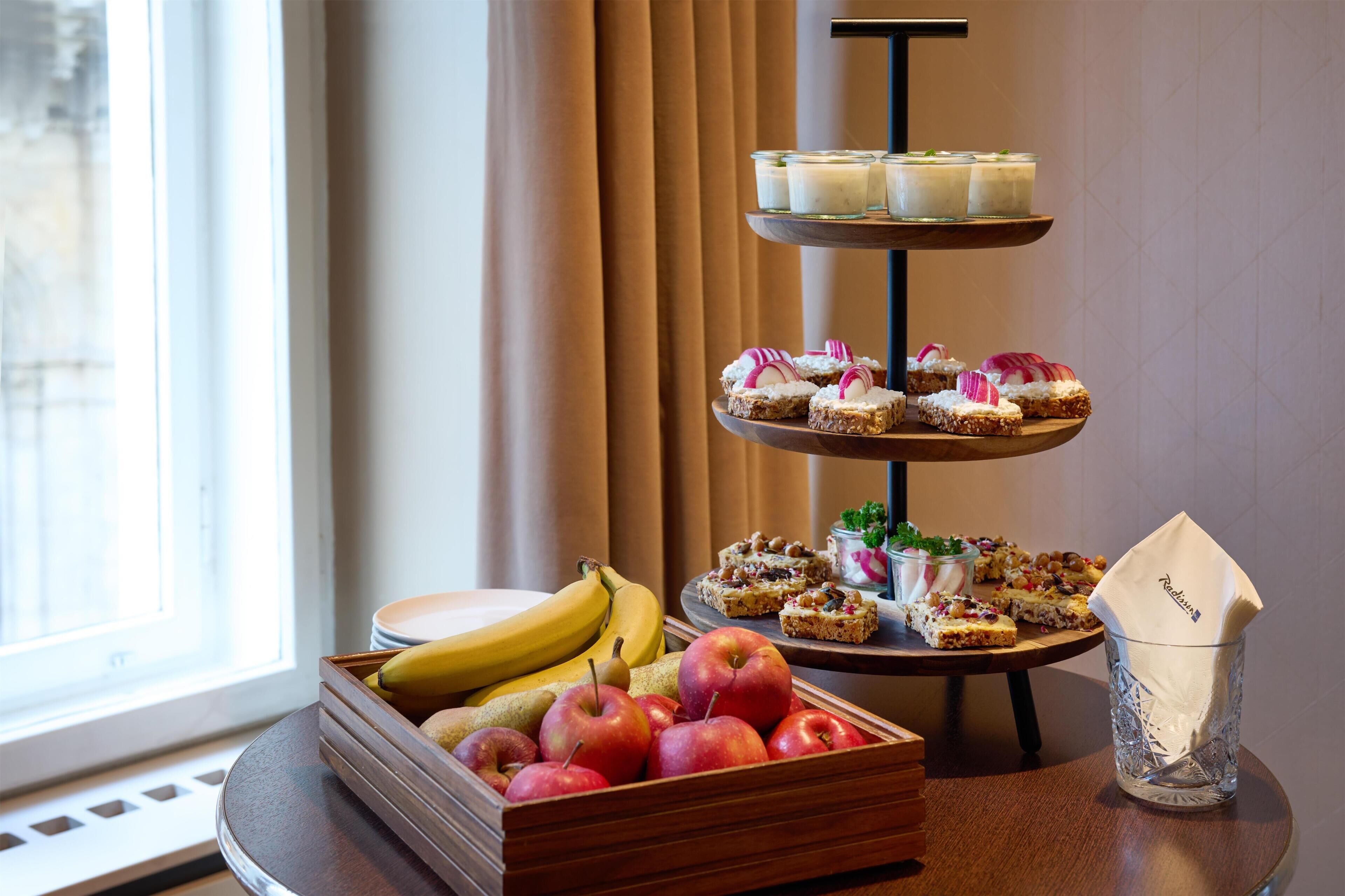 A tiered dessert stand with various pastries, a fruit bowl with bananas and apples, and a glass vase with a napkin.