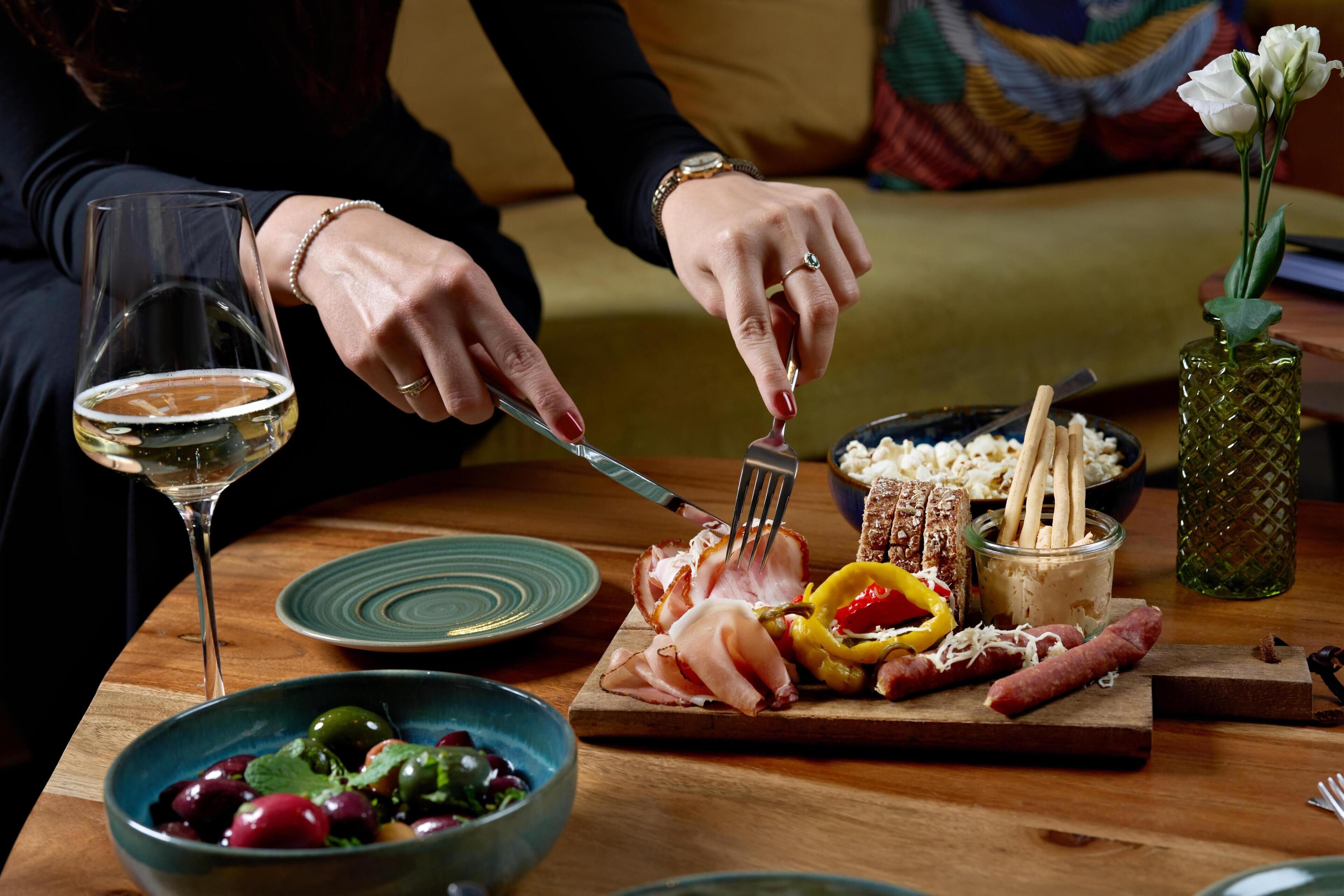 A dining table with a glass of white wine, a bowl of olives, a plate of sliced meats, and a small vase with flowers.
