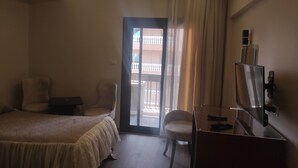 Une chambre d’hôtel avec un lit, un bureau, une chaise et une télévision. Elle dispose d’un balcon avec une balustrade et offre une vue sur l’extérieur.