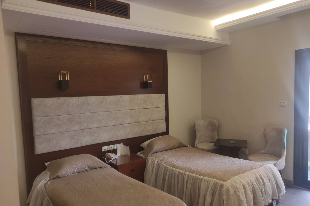 A hotel room with two beds, a wooden headboard, bedside tables, a chair, and a window.