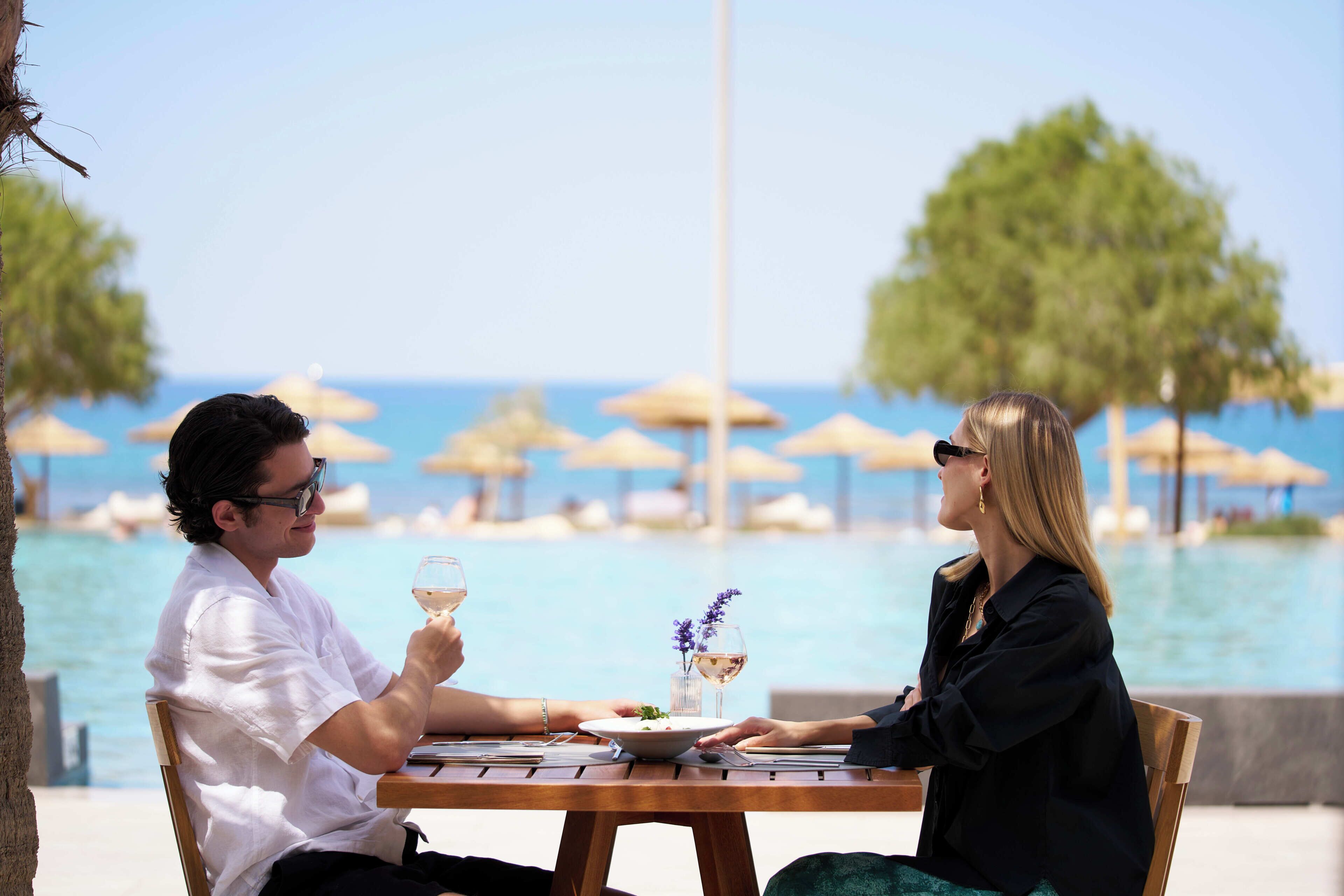 Two people seated at a wooden table with a view of the sea and umbrellas in the background.