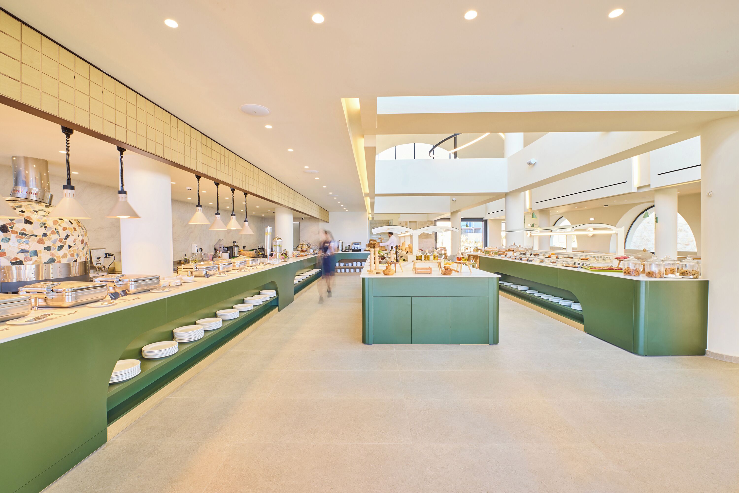 Un vaste espace de restauration proposant de nombreux comptoirs de service et une île centrale, avec un design moderne agrémenté de touches de vert et d’un éclairage abondant.