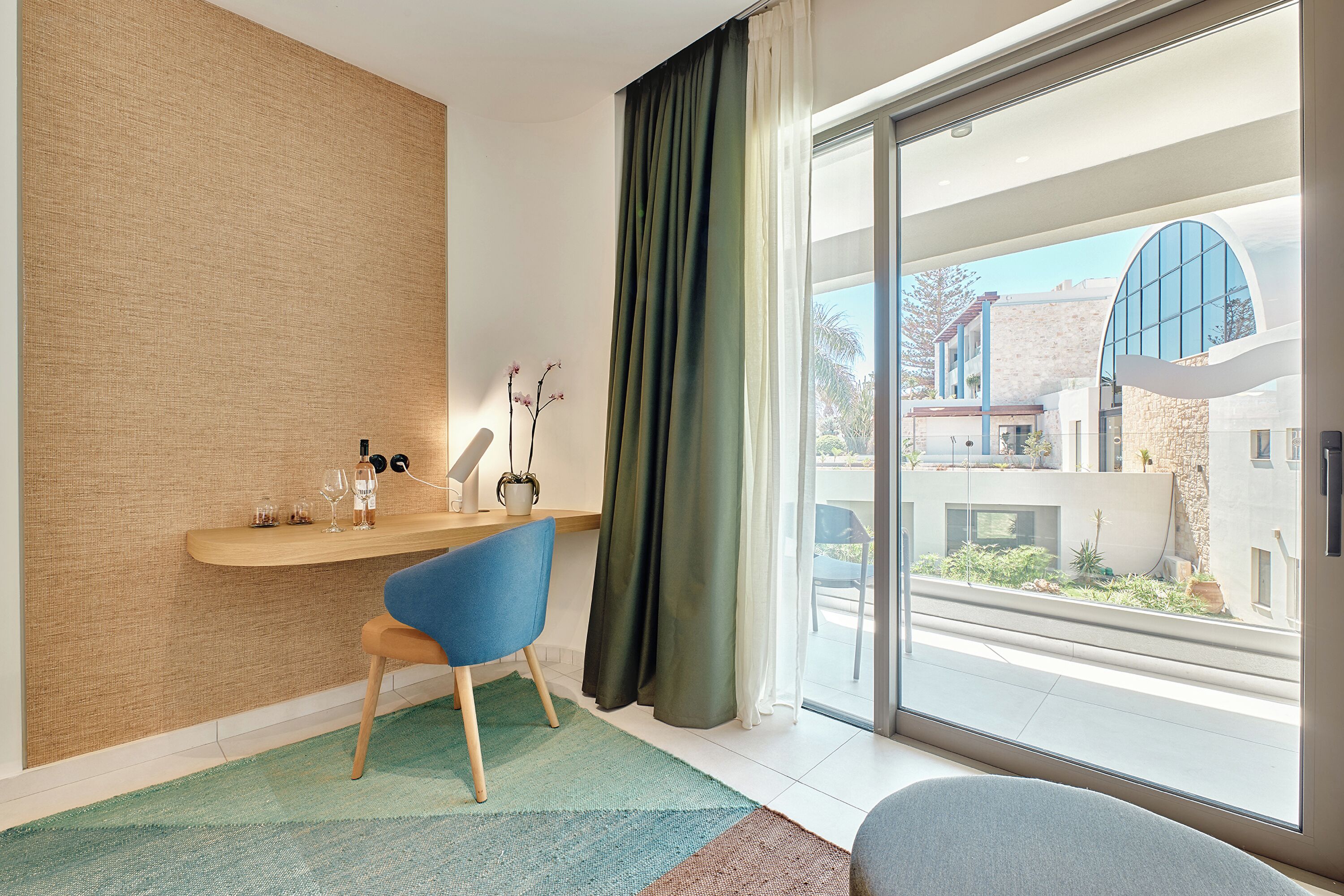 Quarto moderno com escrivaninha de madeira, cadeira azul, janela grande com vista para edifícios e cortina verde.