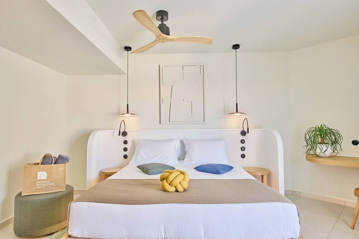 A modern bedroom with a bed, a ceiling fan, bedside lamps, a bedside table, a plant, and a bag of toiletries.
