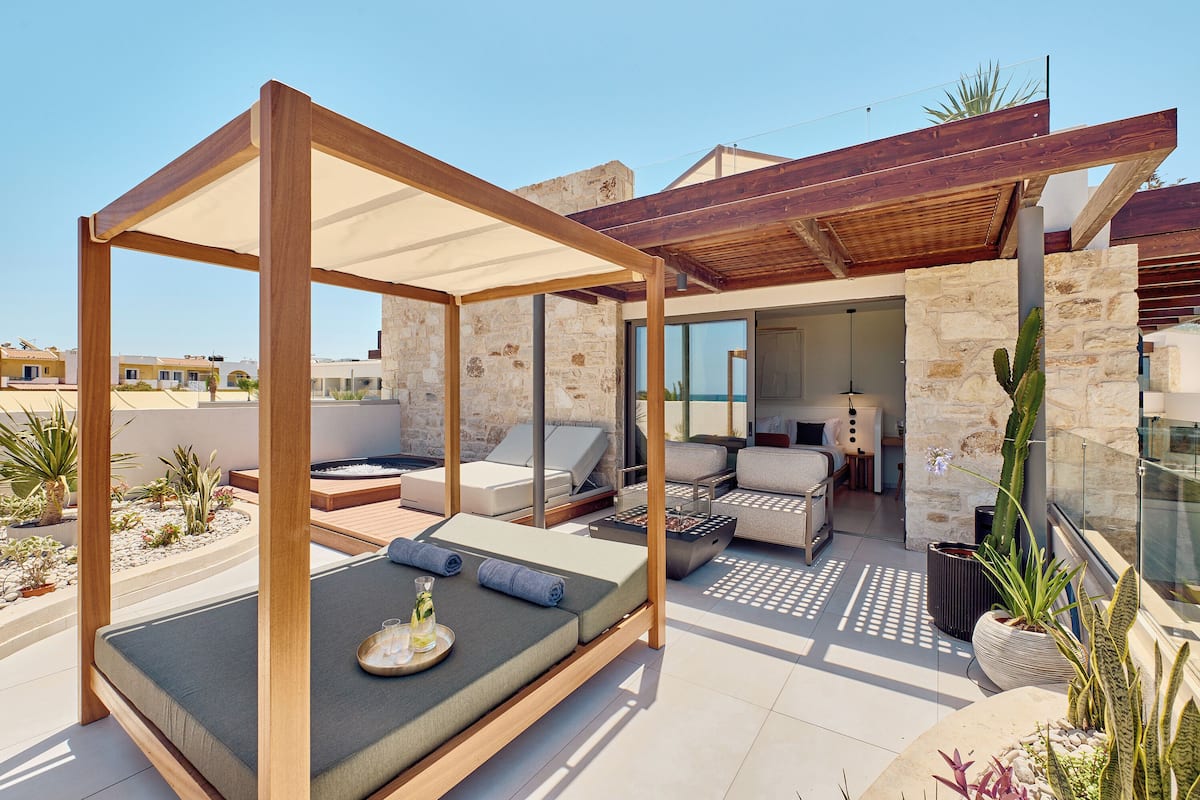 A modern outdoor lounge area with a wooden pergola, cushioned seating, and a stone wall backdrop.