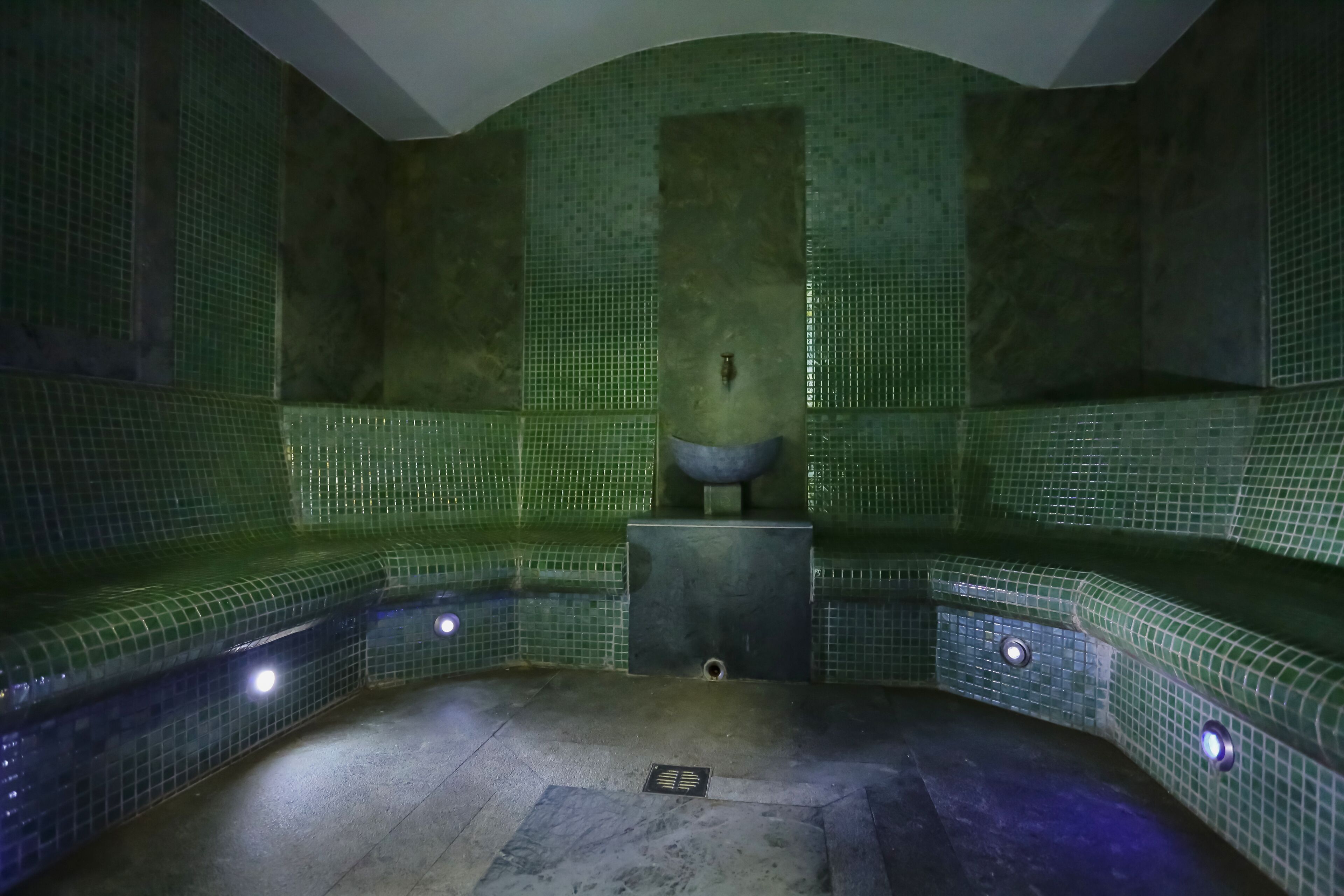 A spa room with green mosaic walls, a central stone basin, and a tiled floor.