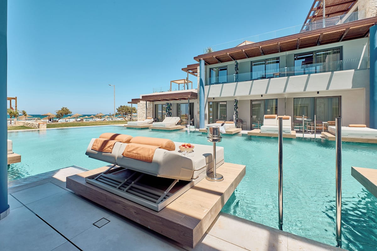 A modern pool area with lounge chairs and a built-in grill, overlooking a clear blue pool and a large building with large windows.