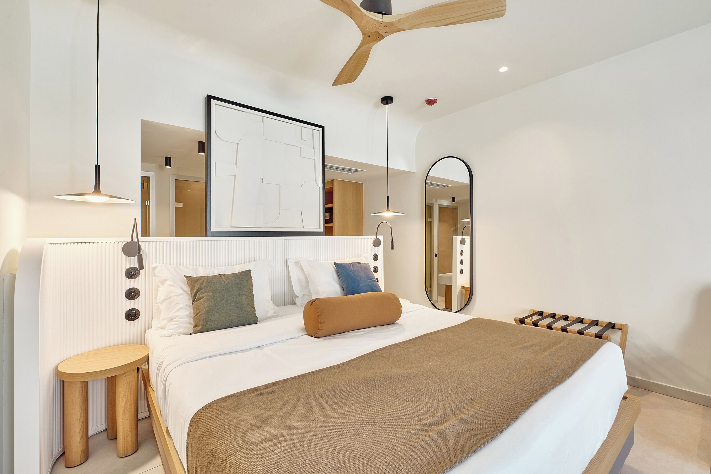 A modern bedroom with a large bed, a round wooden side table, a ceiling fan, and a large mirror.