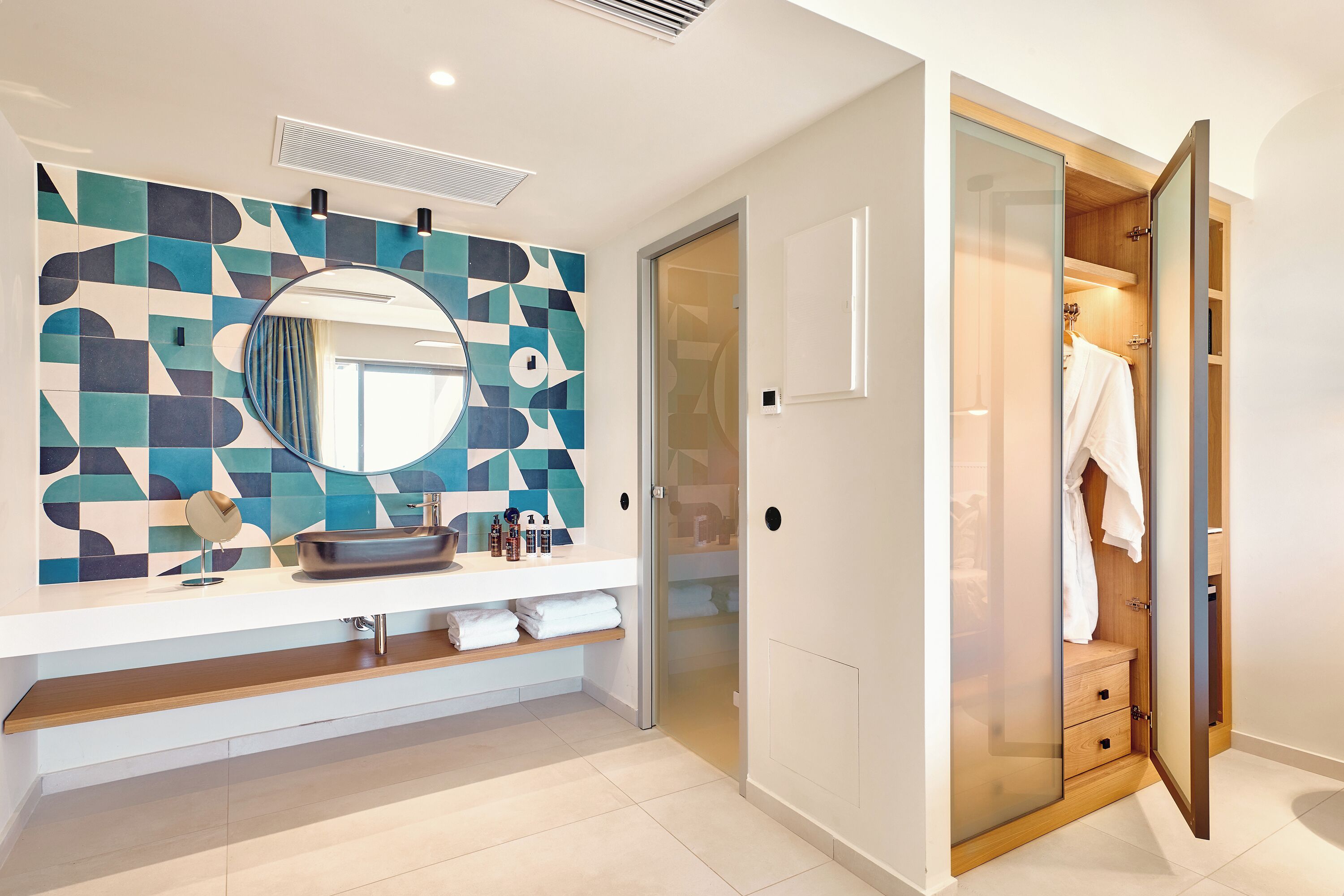 A modern bathroom with a geometric tile backsplash, a round mirror, a built-in sink, and a walk-in closet with sliding doors.