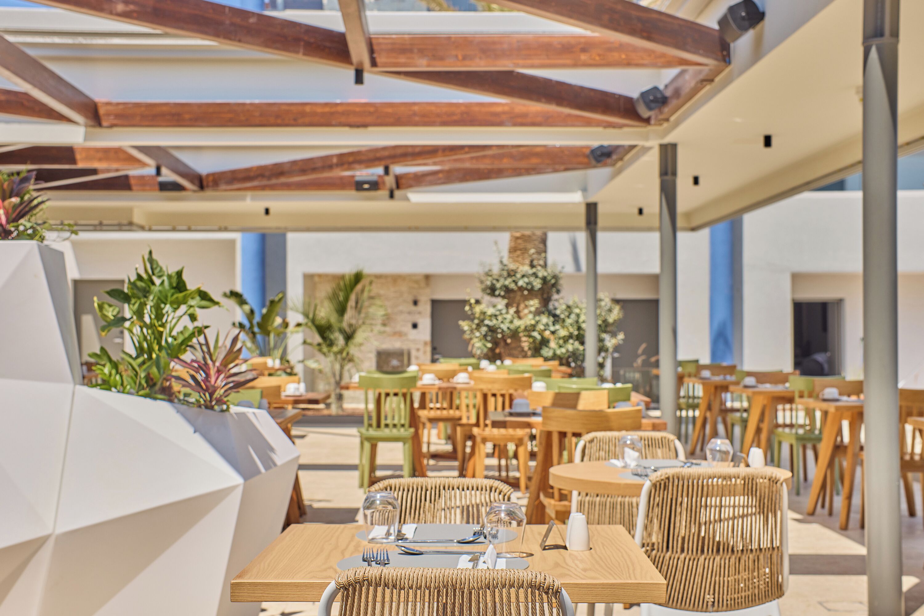 Un espacio exterior para comer con mesas de madera y sillas de mimbre, rodeado de plantas en macetas y un muro de piedra.