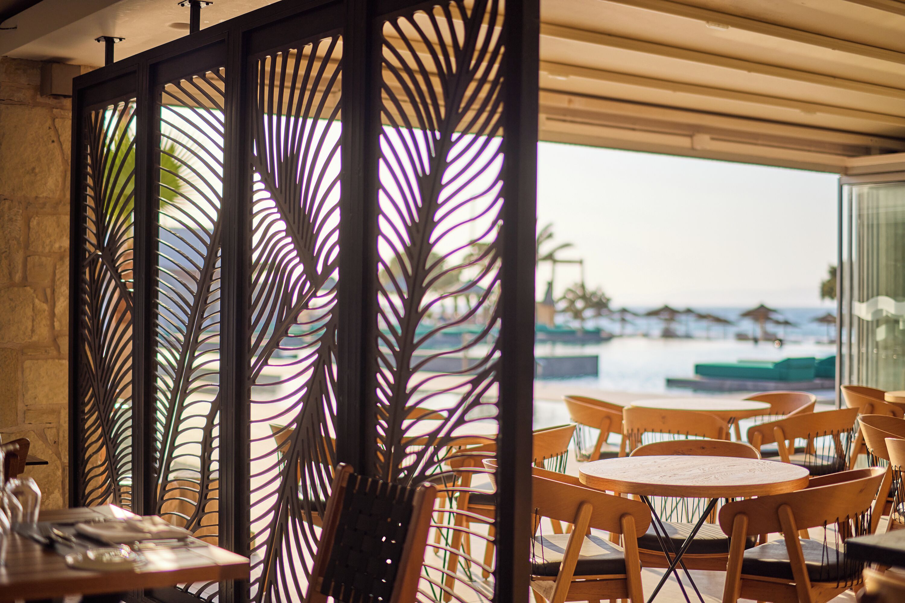 Un restaurante con mesas y sillas de madera, un separador metálico decorativo y vistas al océano y sombrillas.