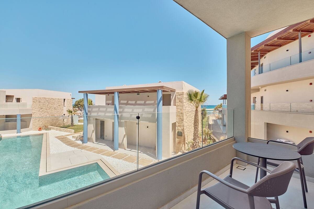 A balcony with a pool, a table, and chairs, overlooking a pool area and buildings.