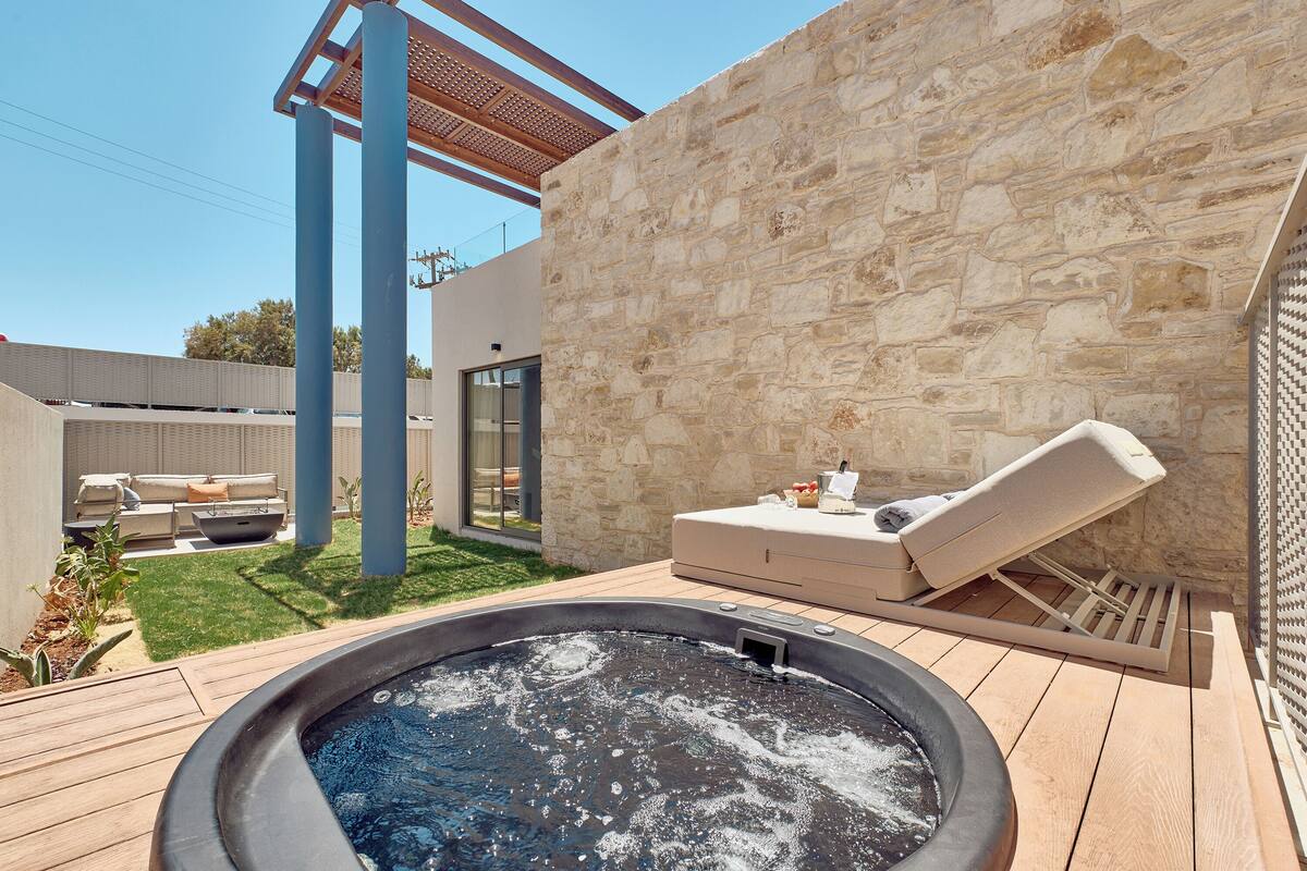 A hot tub in a backyard with a stone wall, a lounge chair, and a patio area.