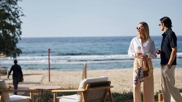Escena en la playa con una silla de madera y una mesa, una persona con un vestido a rayas y otra con una camisa oscura.