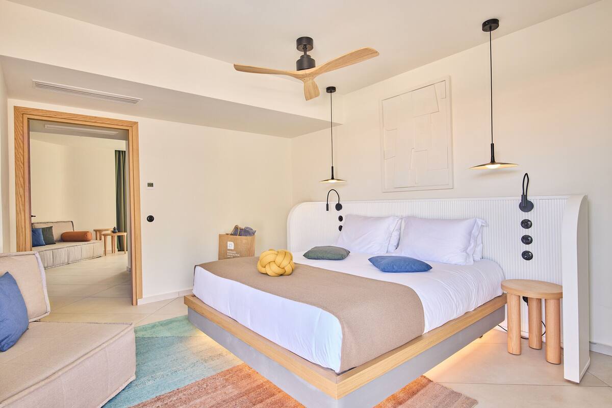 A modern bedroom with a bed, bedside tables, a ceiling fan, and a view into another room.