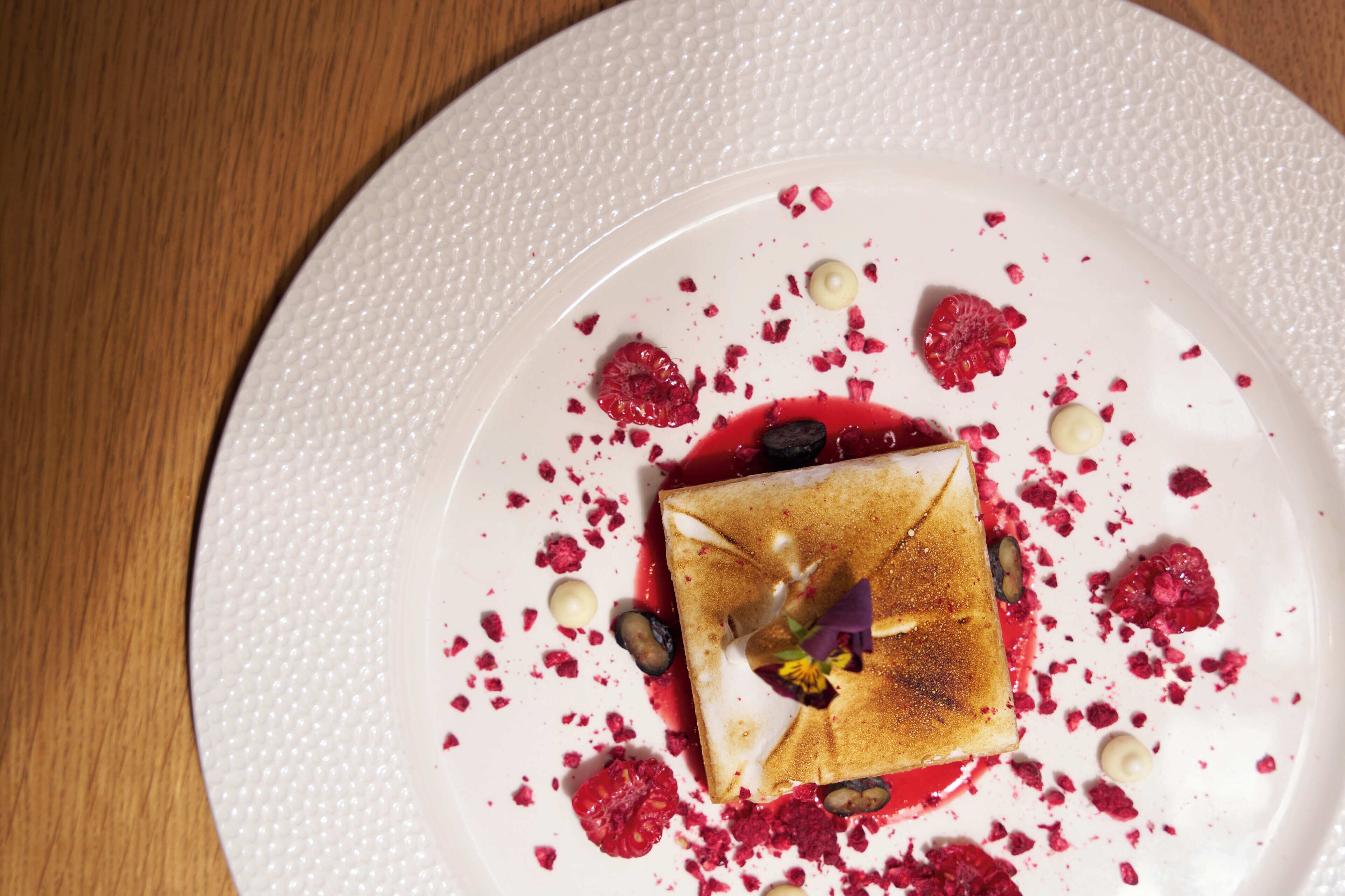 A dessert on a white plate with raspberry pieces and a small flower garnish.