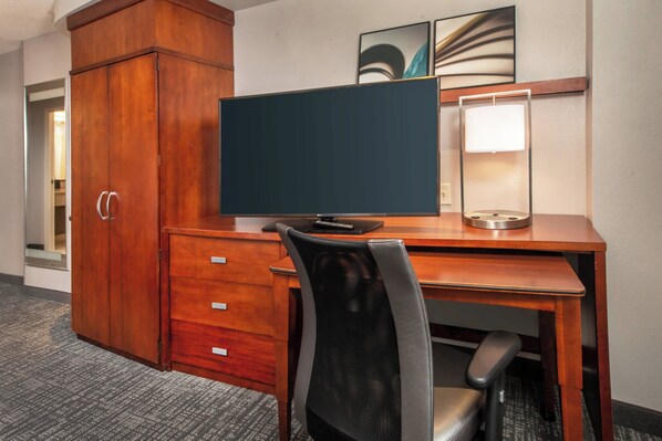 A hotel room with a wooden desk, a computer monitor, a chair, a wardrobe, and a lamp.