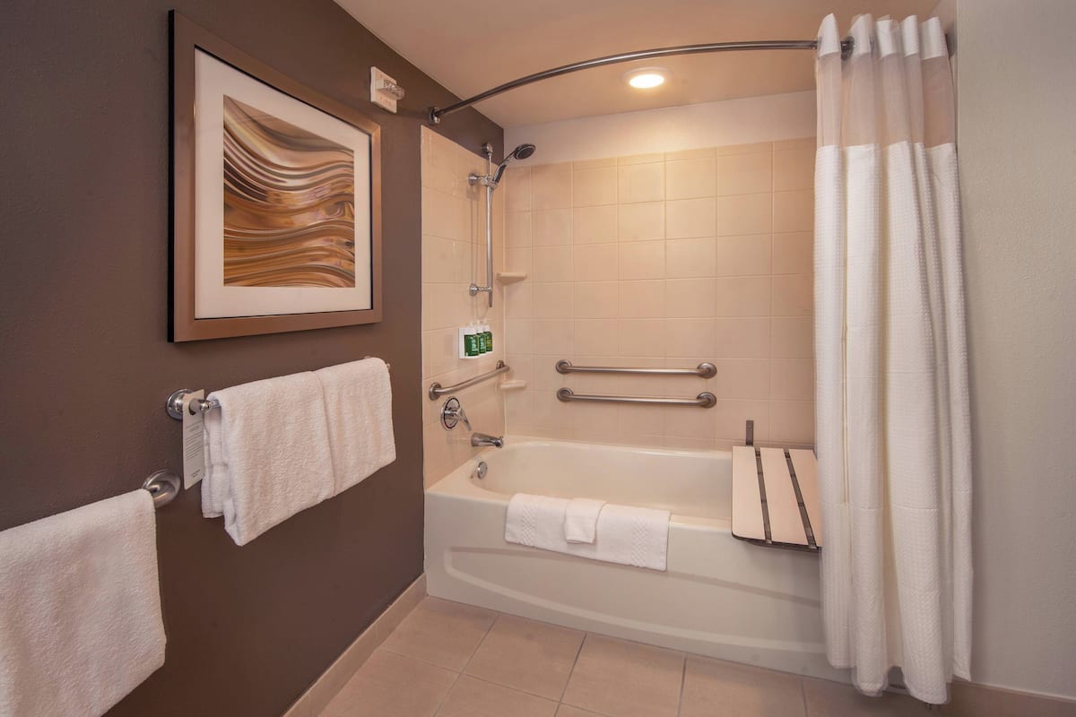 Ein modernes Badezimmer mit Badewanne, Dusche und Toilette. Zwei Handtücher hängen an Haken, ein gerahmtes Kunstwerk ist an der Wand angebracht, und ein weißer Duschvorhang ist vorhanden.