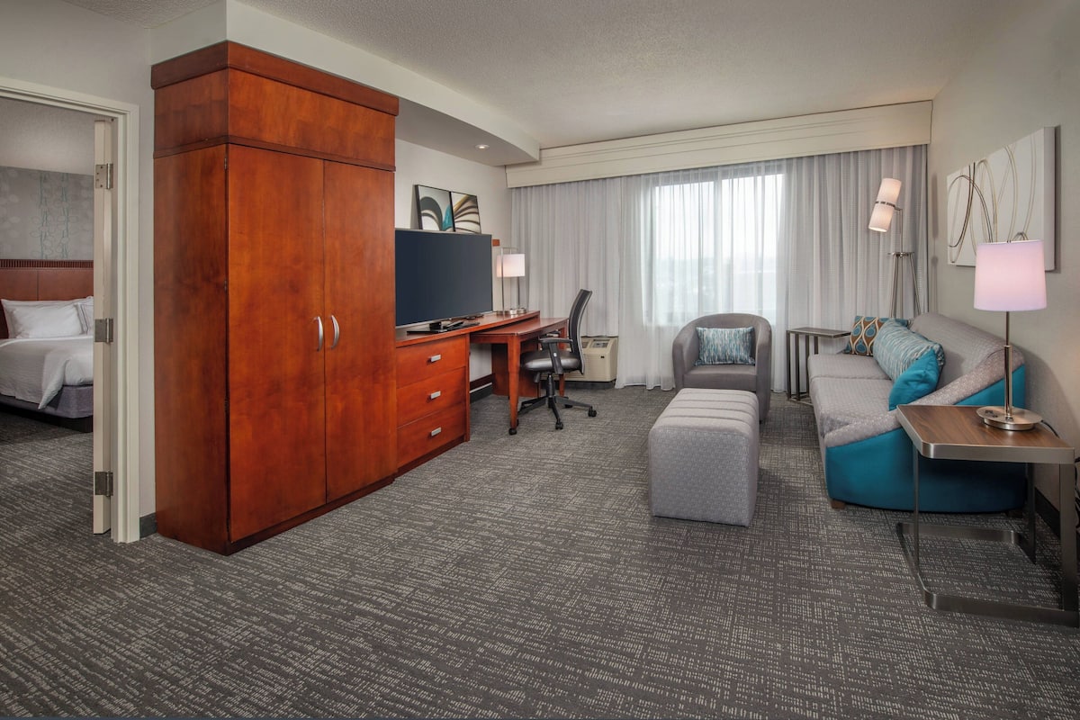 Ein Hotelzimmer mit Holzschrank, Schreibtisch mit Stuhl, grauem Sofa, kleinem Tisch und Fernseher an der Wand.
