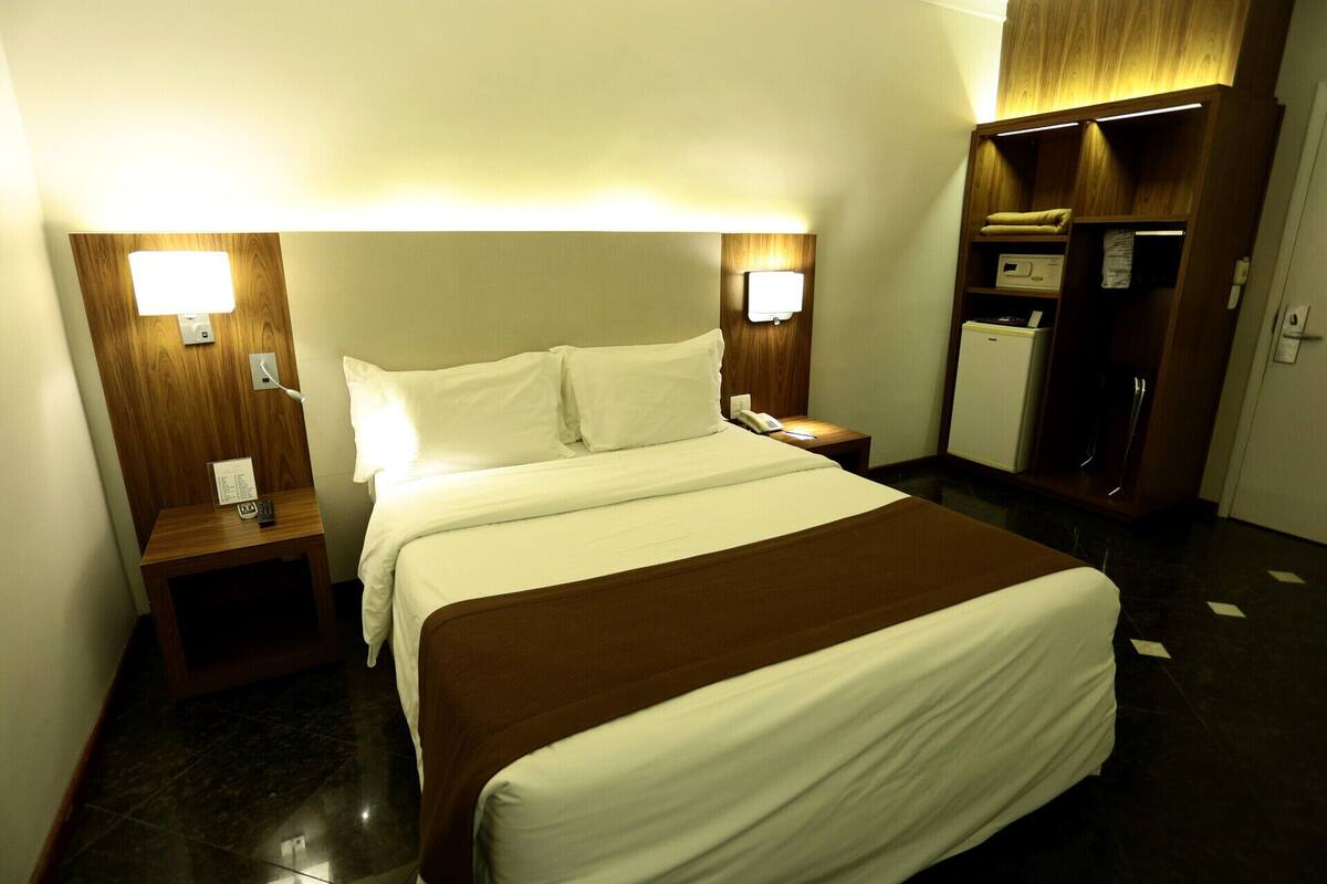 A hotel room with a bed, bedside tables, a nightstand, a wardrobe, and a small refrigerator.