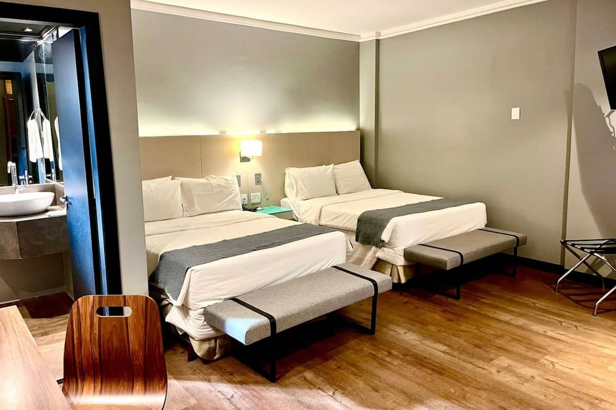 A hotel room with two beds, a bedside table, a mirror, a sink, and a chair.