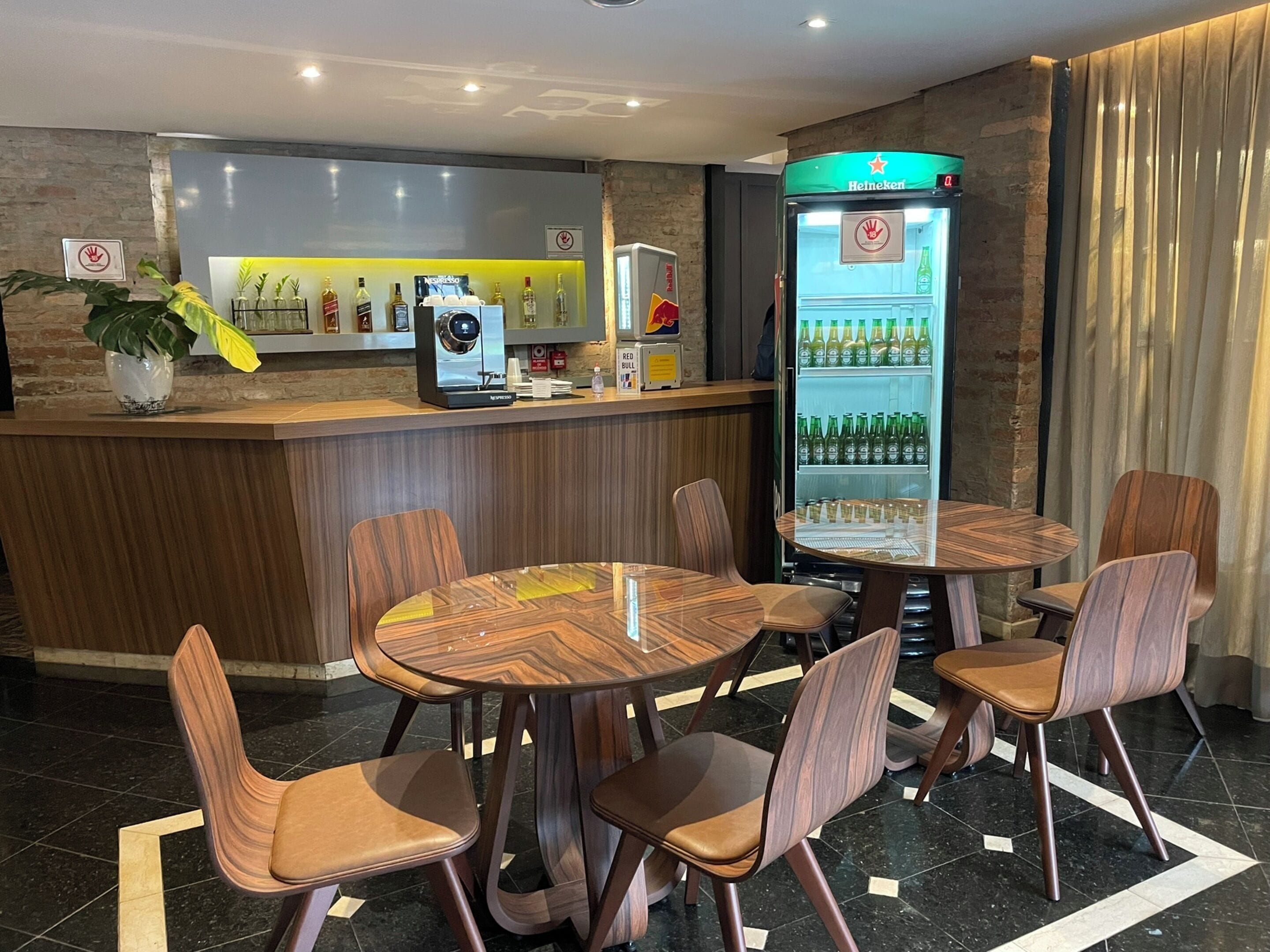 A hotel lobby with a reception desk, a coffee machine, a beverage dispenser, and a seating area with round tables and chairs.