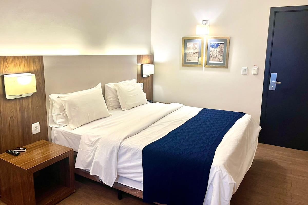 A hotel room with a bed, bedside table, lamp, wall-mounted light, and framed pictures.