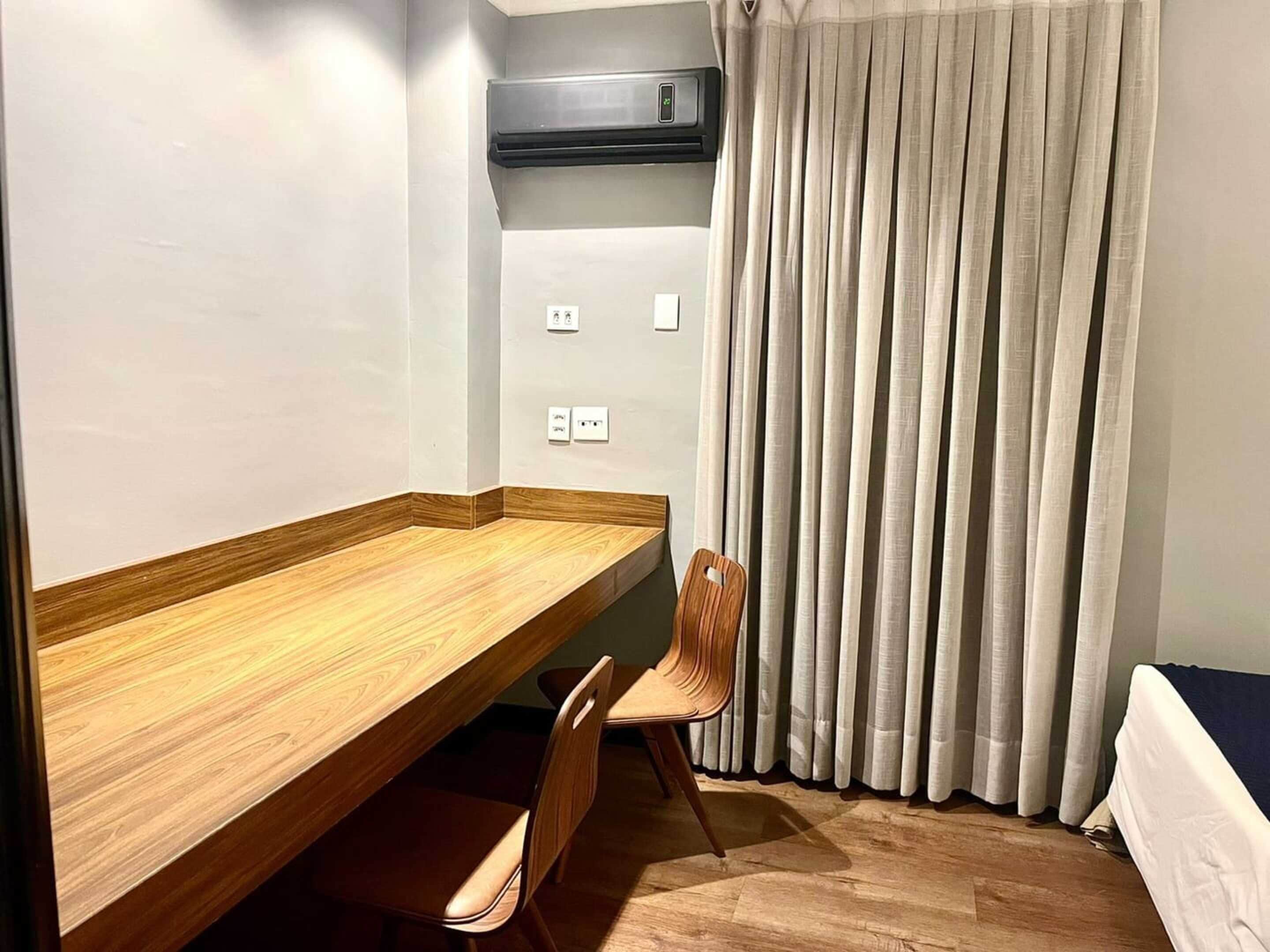 A room with a wooden desk, two chairs, a bed, a window with curtains, an air conditioning unit, and electrical outlets.
