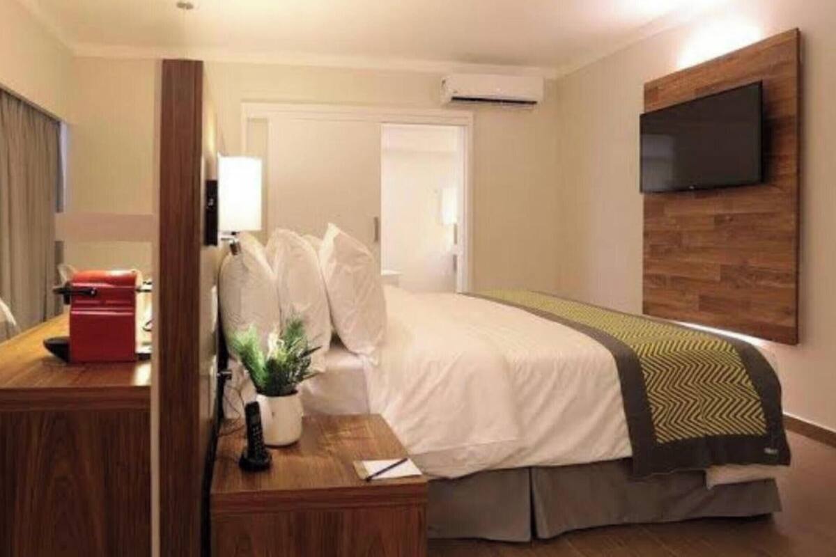 A hotel room with a bed, a desk, a chair, a TV mounted on the wall, and an air conditioning unit.
