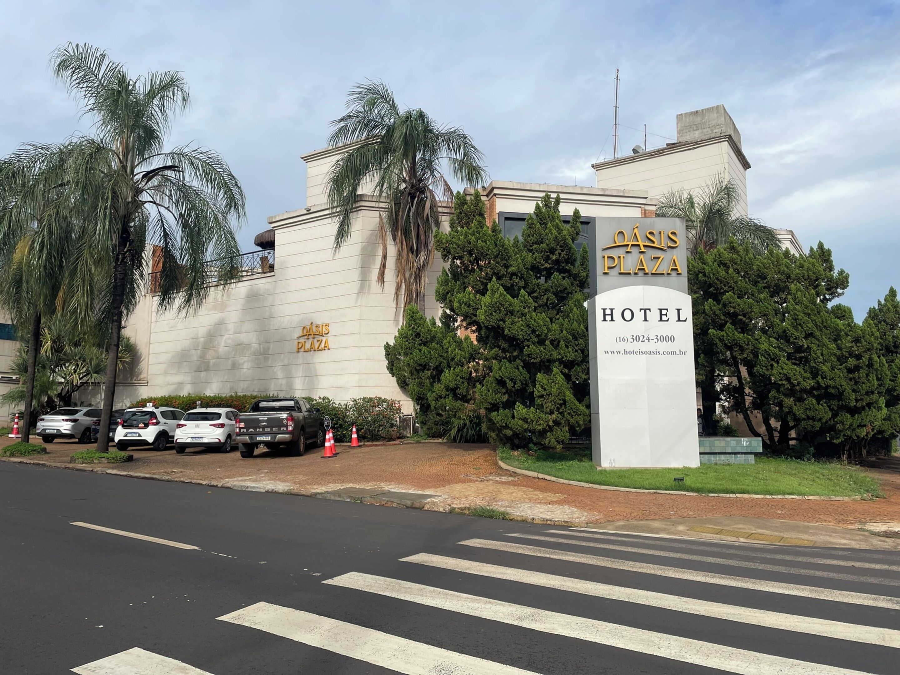 Entrada de un hotel con un cartel que dice 'OASIS PLAZA HOTEL', un estacionamiento con varios autos y palmeras.