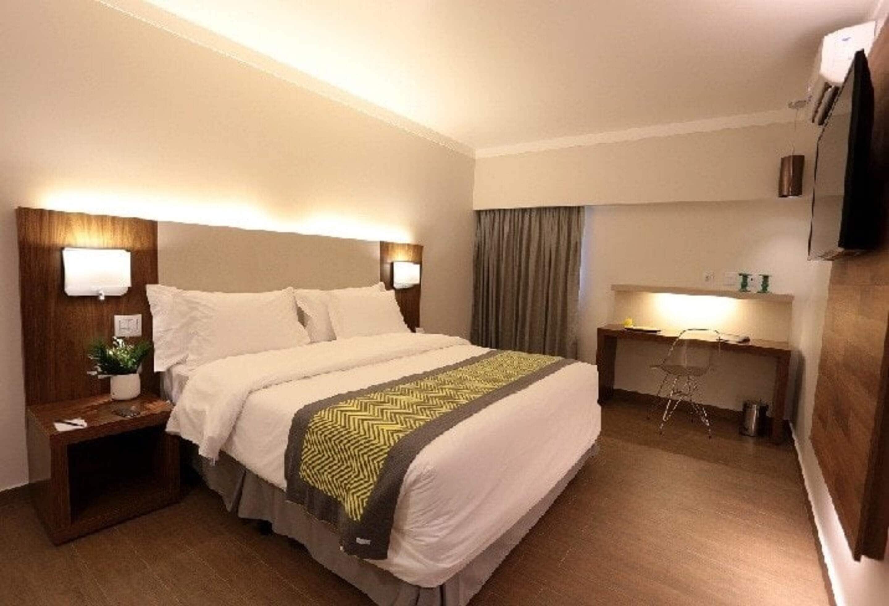 Quarto de hotel com cama, mesas de cabeceira, escrivaninha, cadeira e televisão.