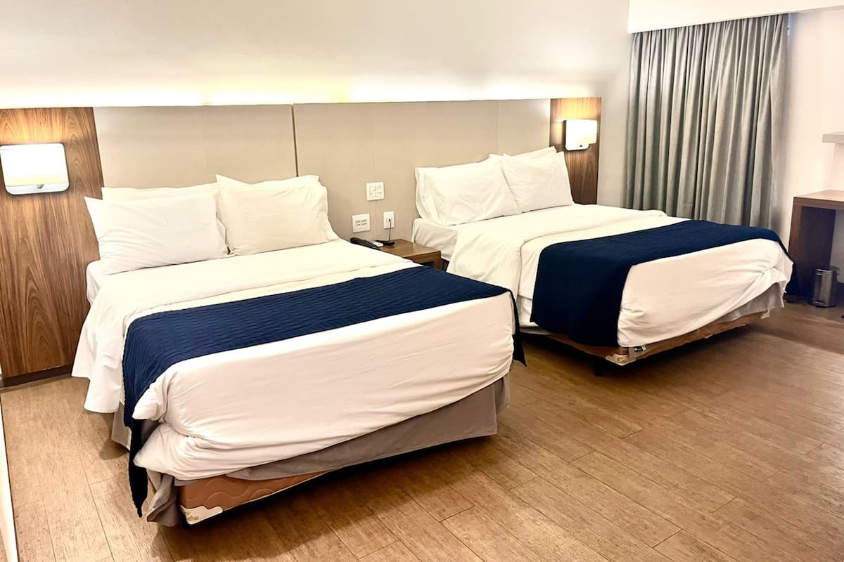 A hotel room with two beds, wooden flooring, a headboard with a light fixture, and a curtain on the window.