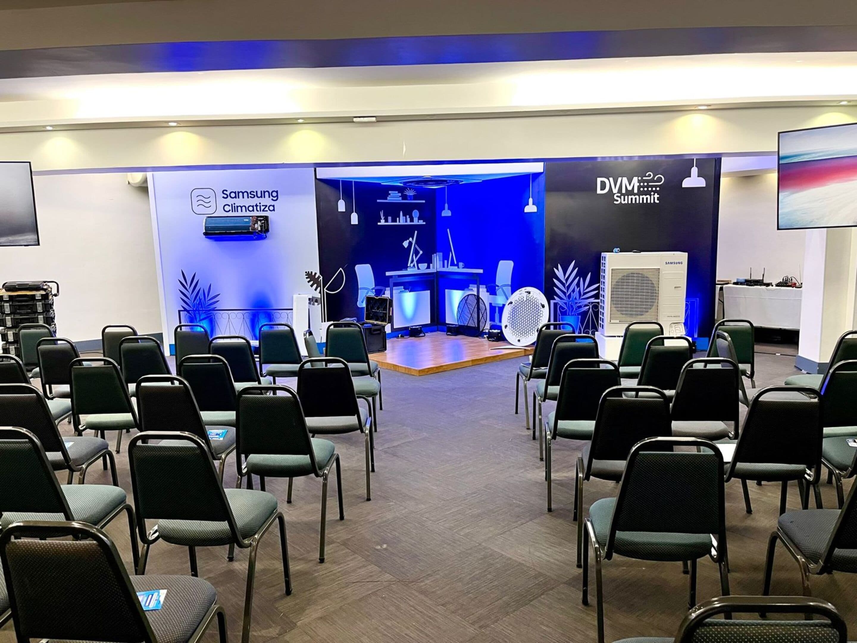 Una sala conferenze con file di sedie, un condizionatore Samsung Climatiza, un display DVM Summit e un grande schermo.