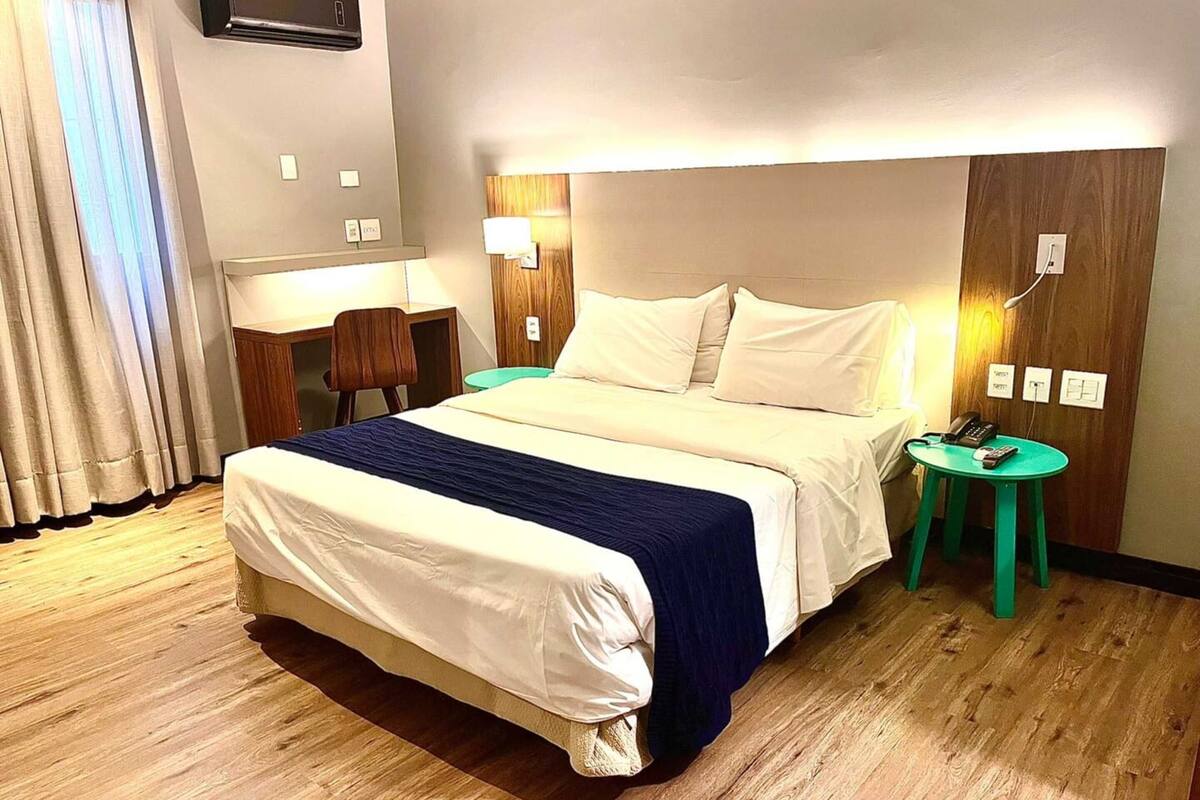 A hotel room with a bed, bedside tables, a desk, a chair, and a green stool.